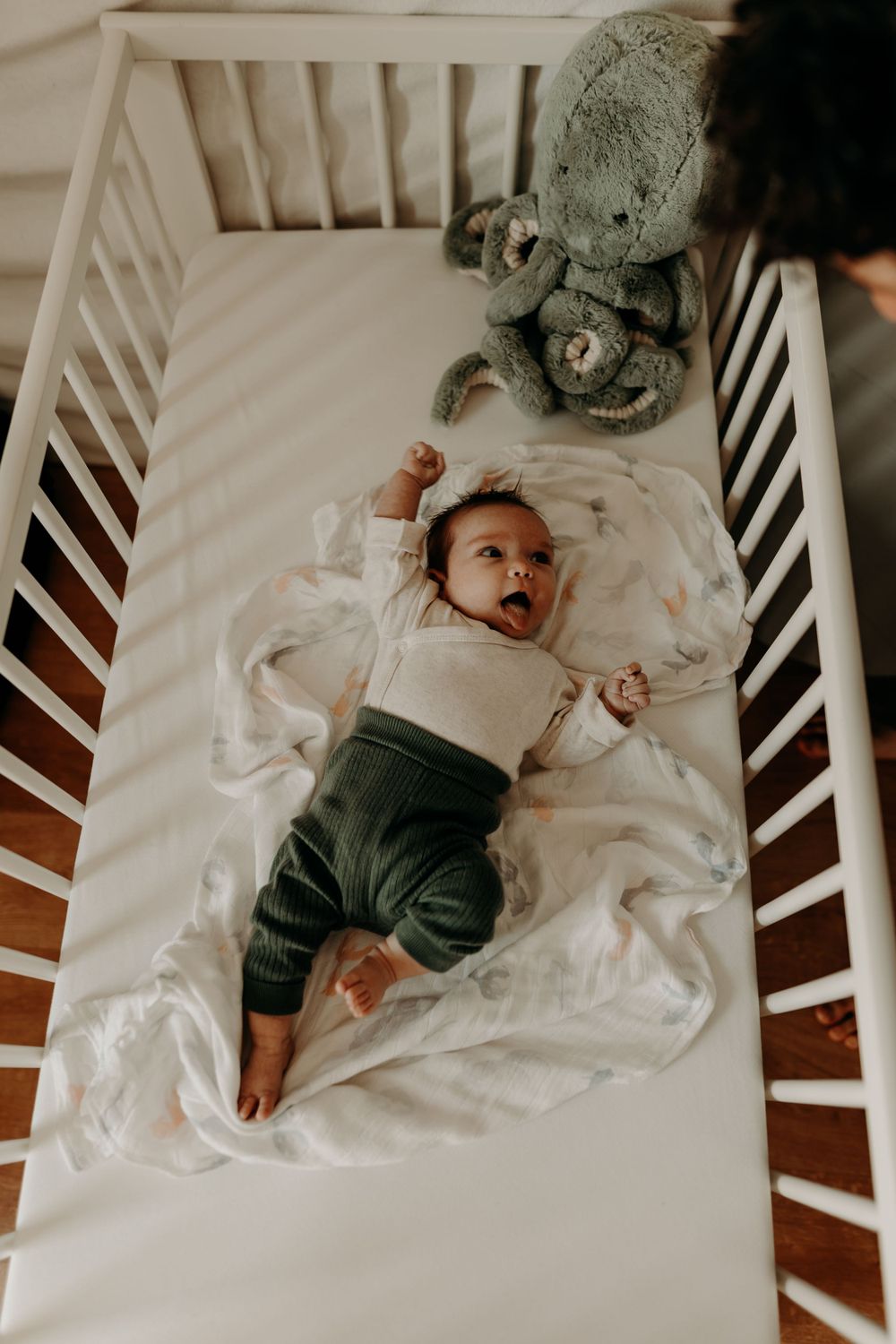 Baby lying in white crib with stuffed elephant toy against white bedding.