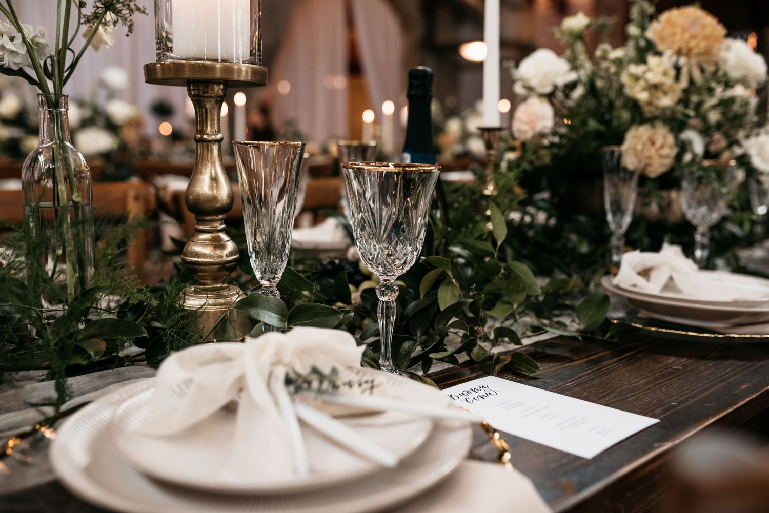 Close-up of sophisticated wedding place setting with crystal glasses, white napkin and greenery accents.