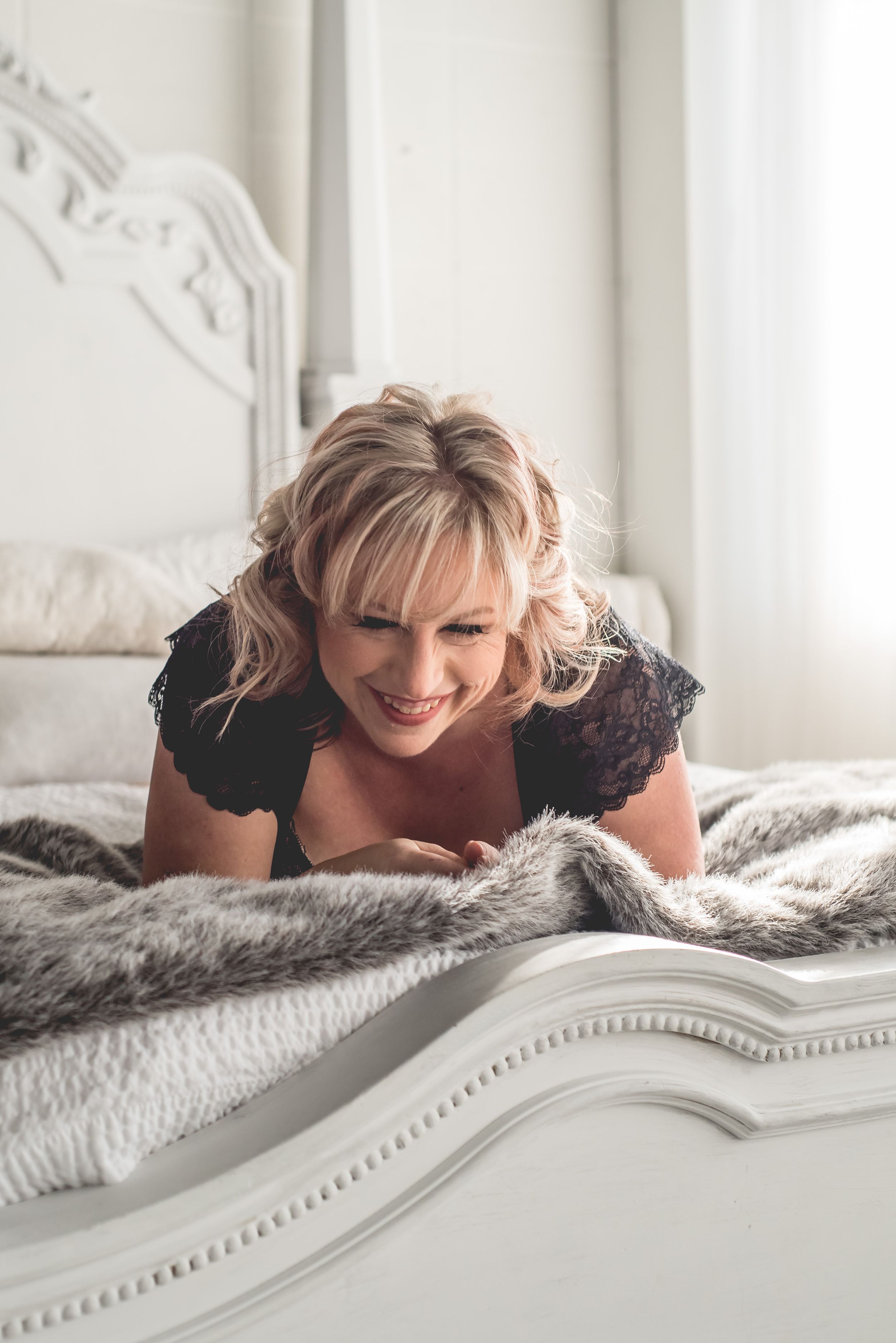 A person in black lace lounges on a white fur blanket while smiling.