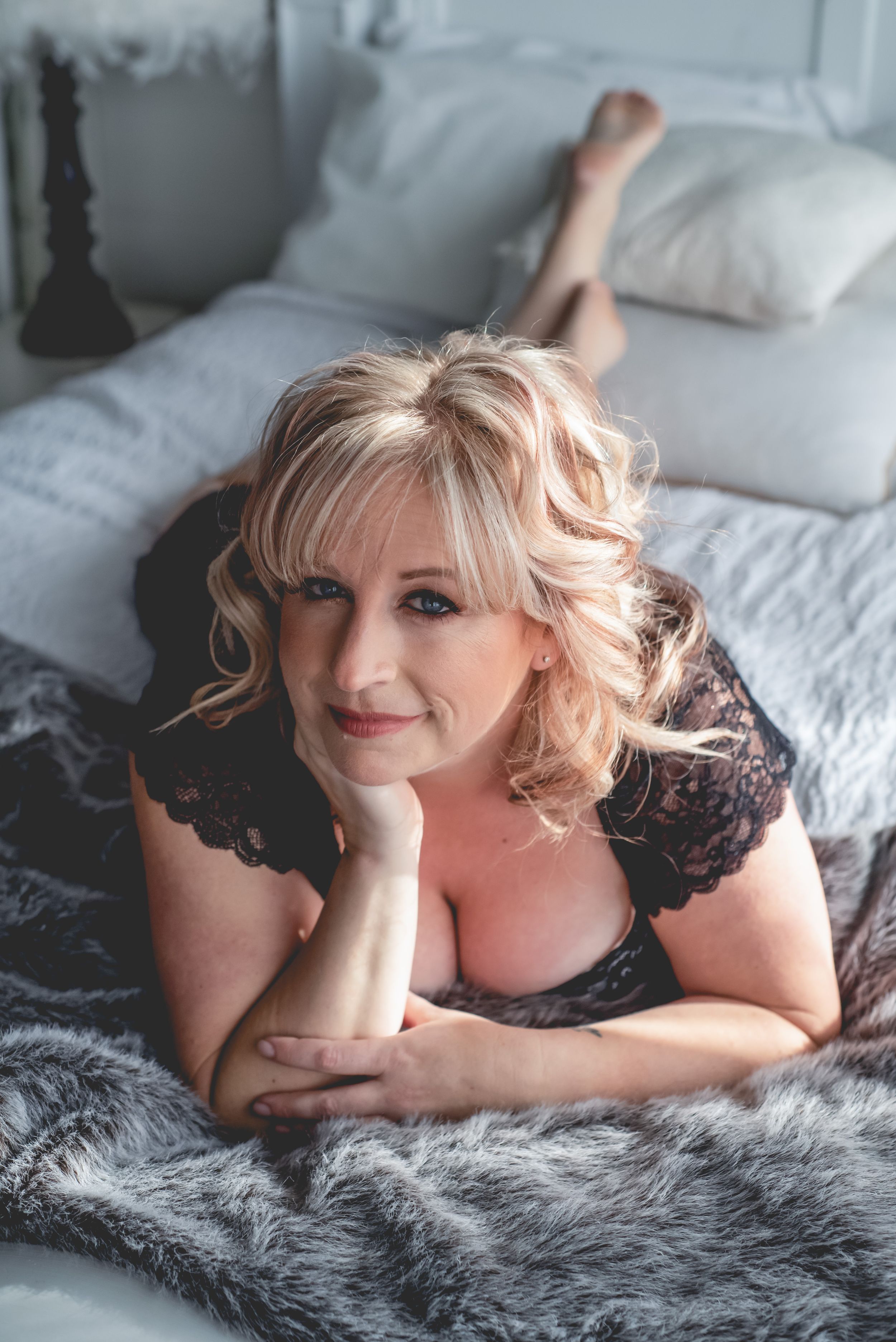 A person in black lace poses on a grey fur blanket with a bright smile.