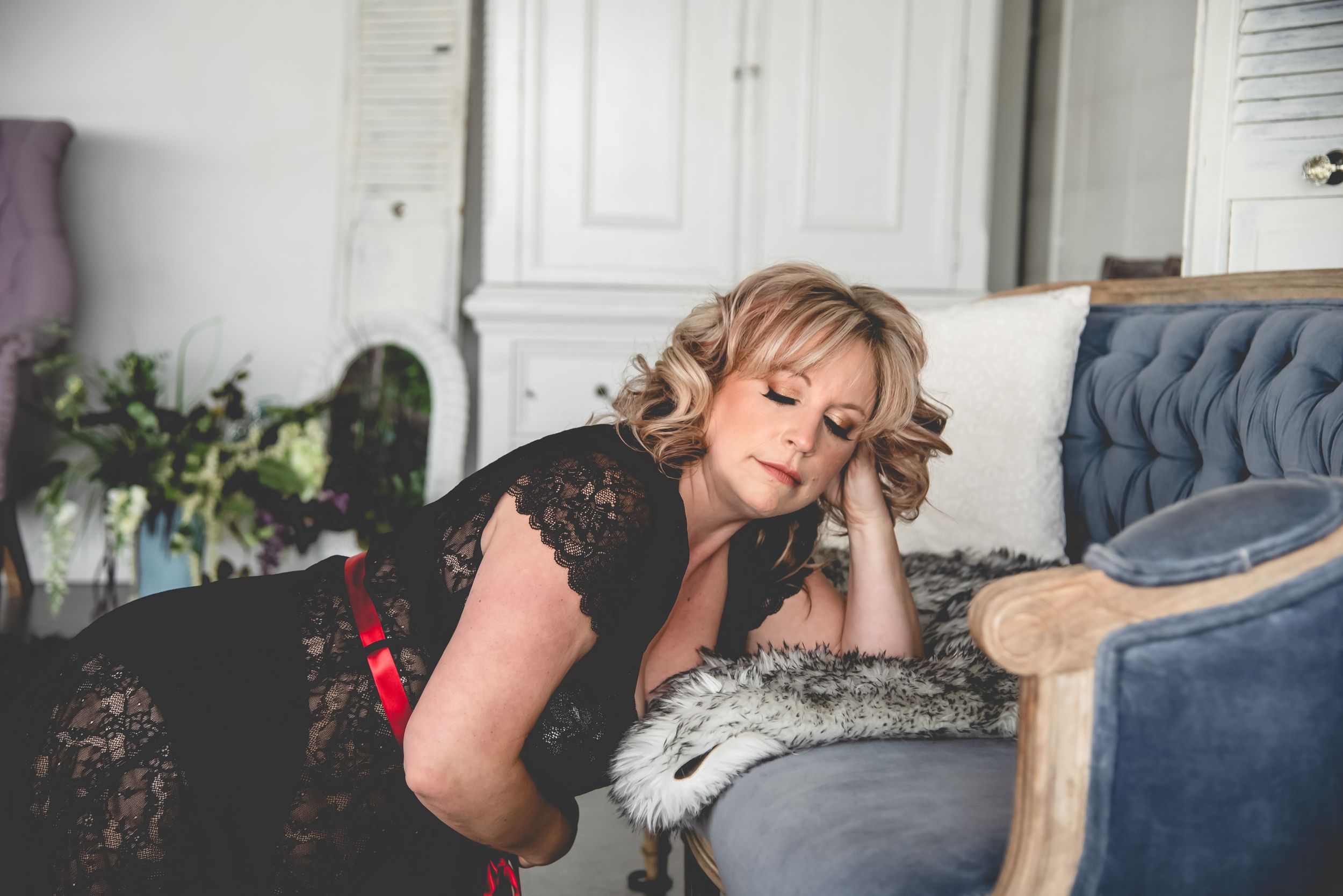 A person in a black lace top poses on a grey couch with decorative pillows.