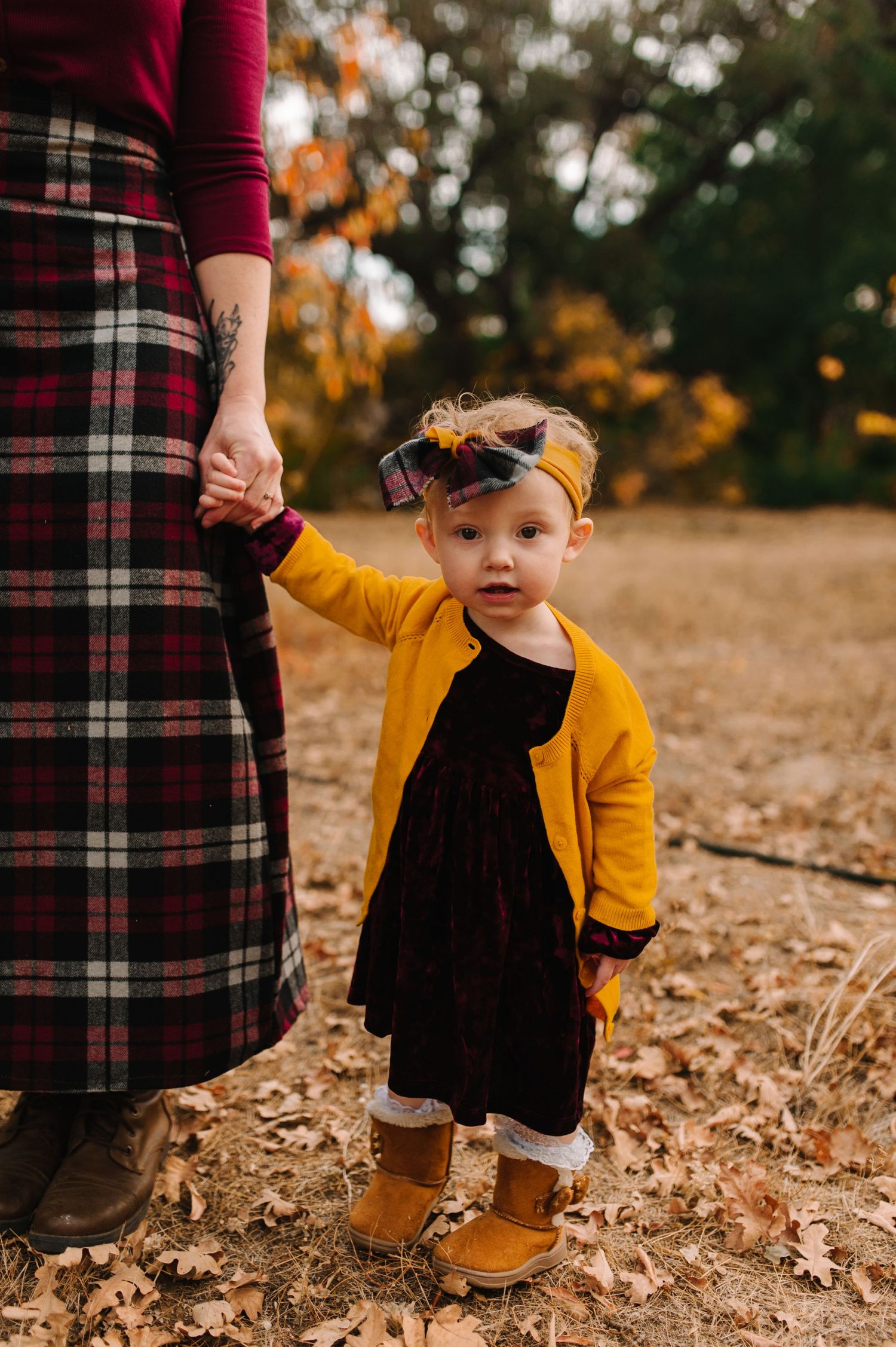 A small child in a yellow cardigan holds hands with someone wearing a plaid skirt on fallen leaves.