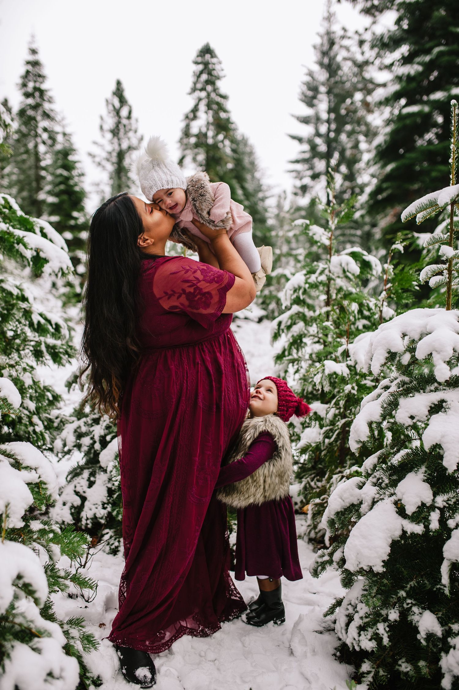 A magical winter scene with burgundy dresses against snow-covered evergreen trees.
