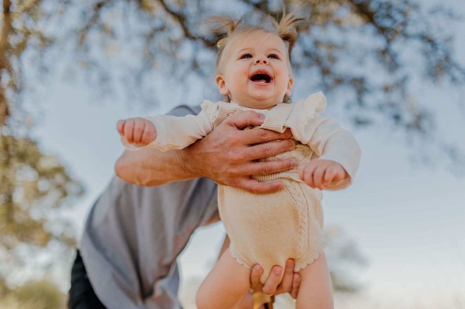 A baby in white clothing is lifted into the air with joyful expression against a blue sky.