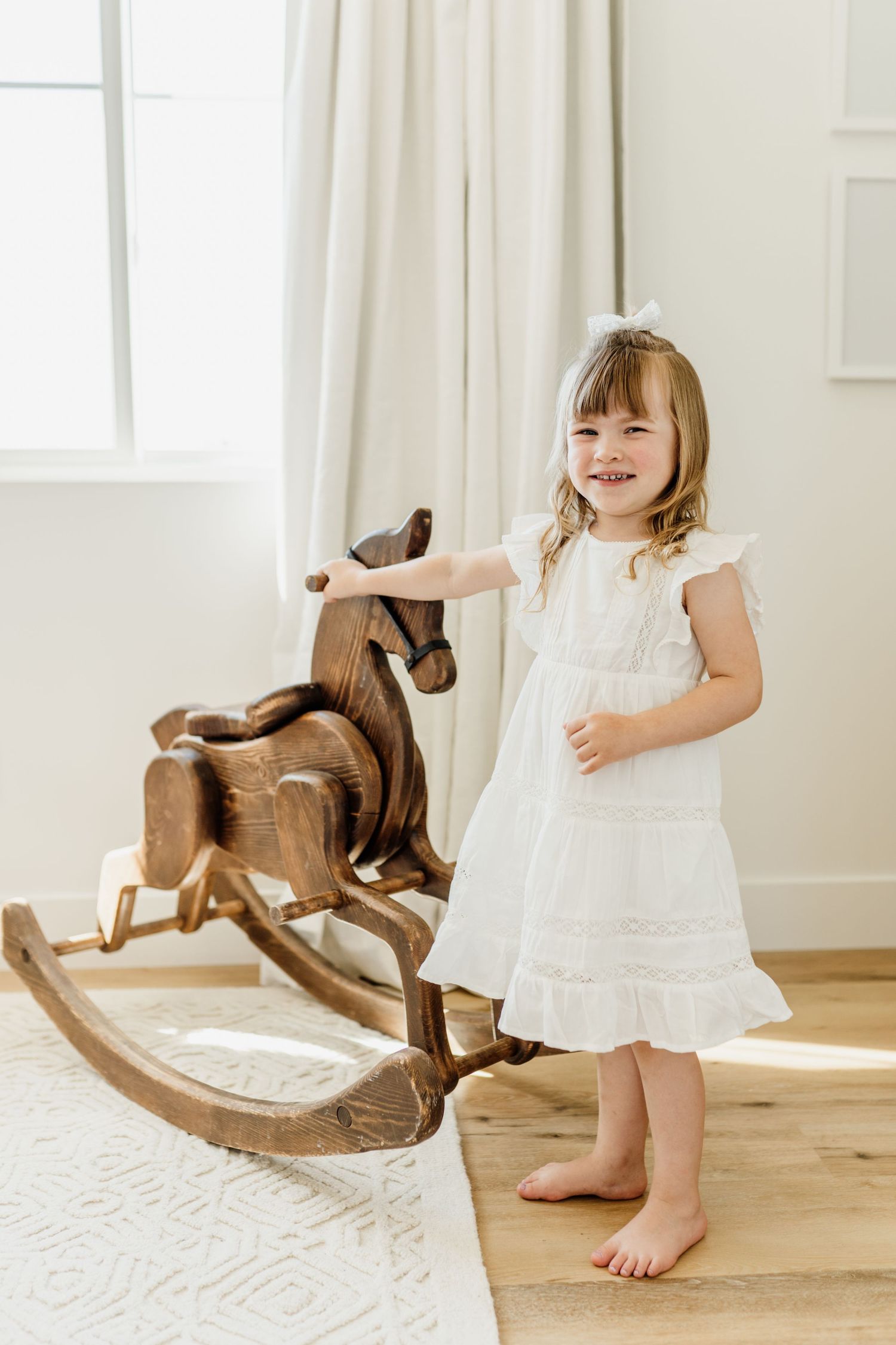 A child in a white dress stands barefoot next to a wooden rocking horse in a bright, airy room.