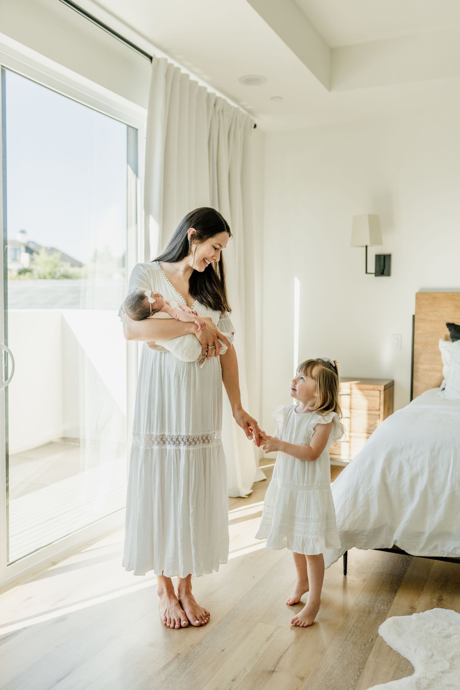 Mother and daughter share a tender moment in a bright white room wearing matching flowy white dresses.