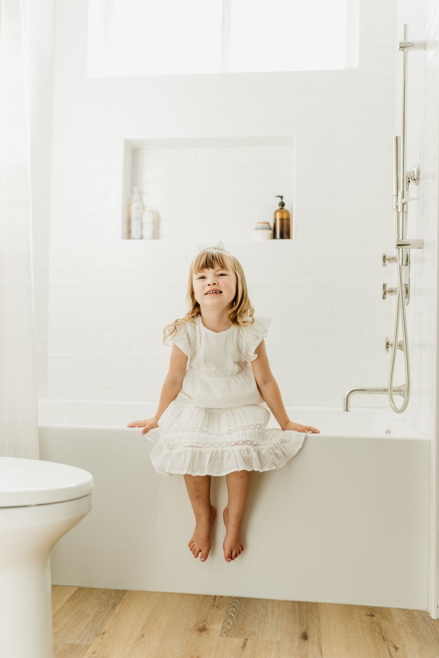 Young child in a white ruffled dress sits on edge of white bathtub in a bright minimal bathroom with wooden floors.