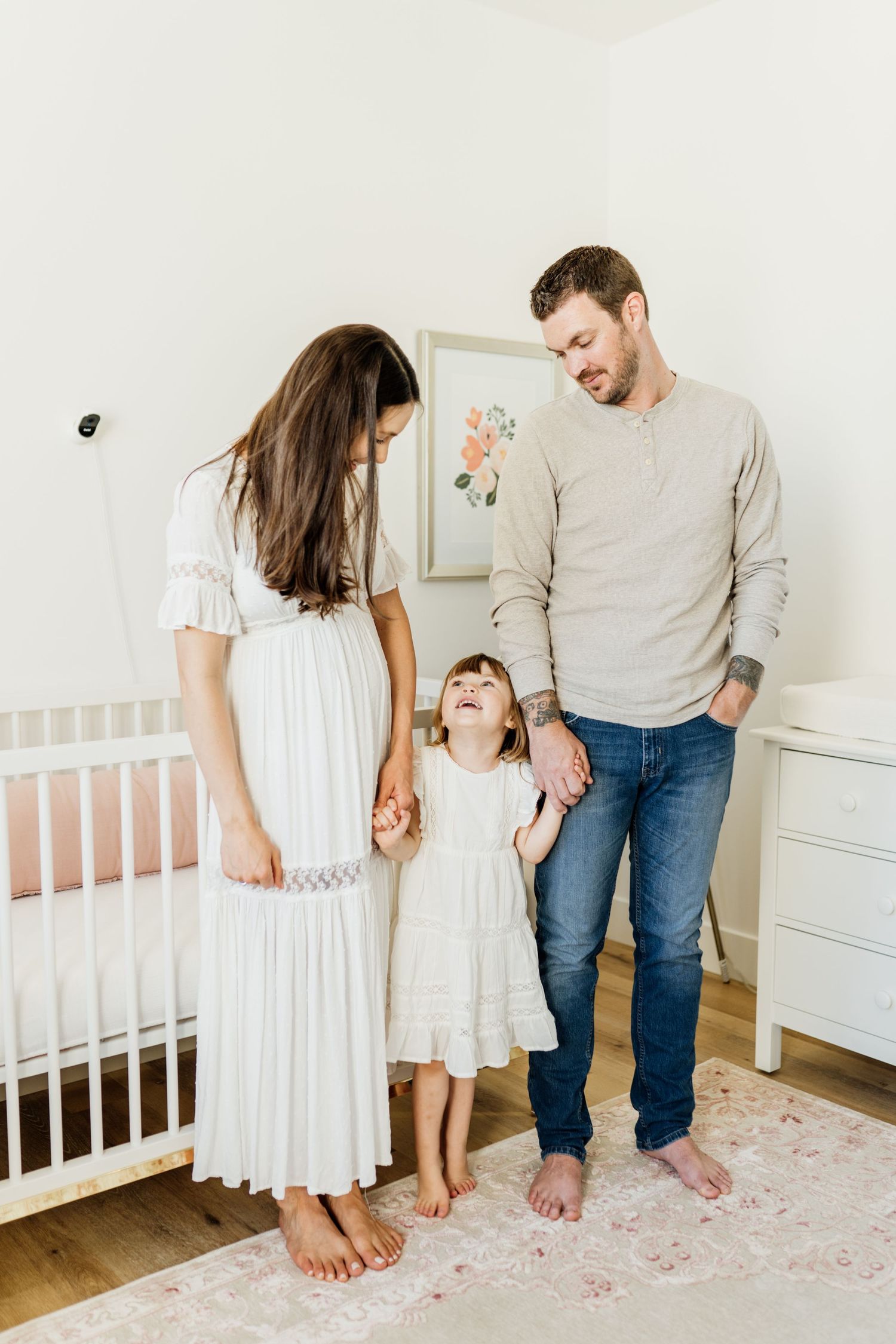 Family stands barefoot together in a bright nursery with white crib and dresser on a floral rug.