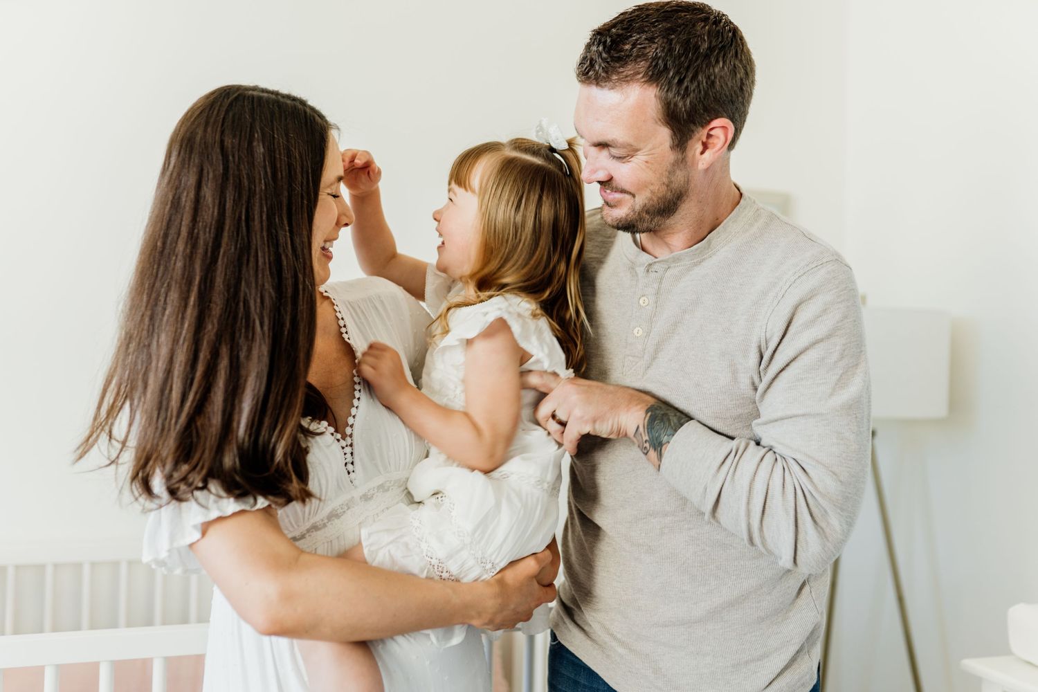 Loving family moment in a bright nursery as parents hold their young child with a white wooden crib in background.