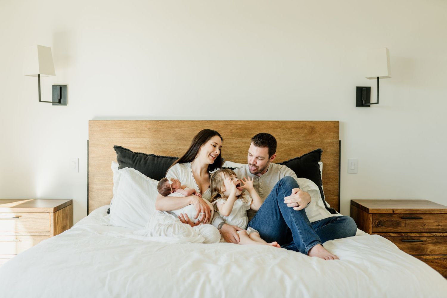 A family lounges together on a white bed with wooden headboard and modern wall sconces in a bright room.
