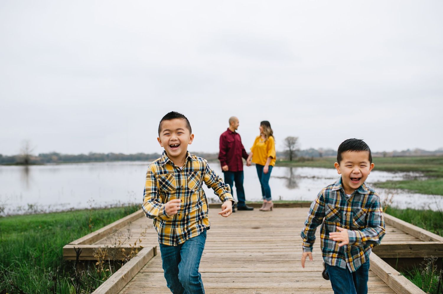 Children run playfully on wooden boardwalk while adults watch in background near a lake.