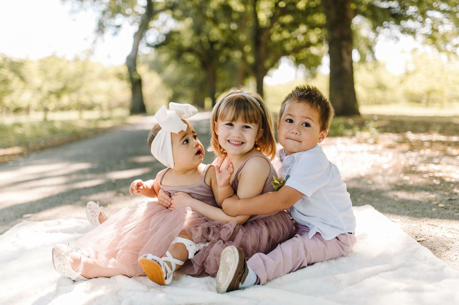 Three young children sitting together on a white blanket outdoors.