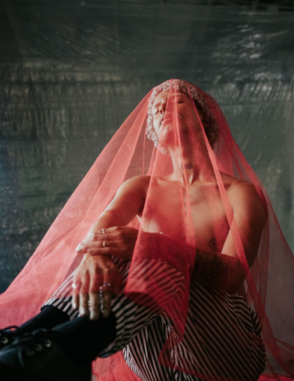 Figure in striped dress draped with flowing red sheer veil in dramatic lighting.