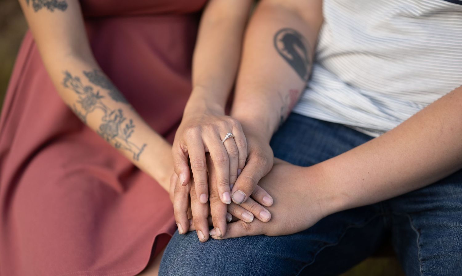 Close-up of hands with tattoos and engagement ring resting together on laps showing intimacy.