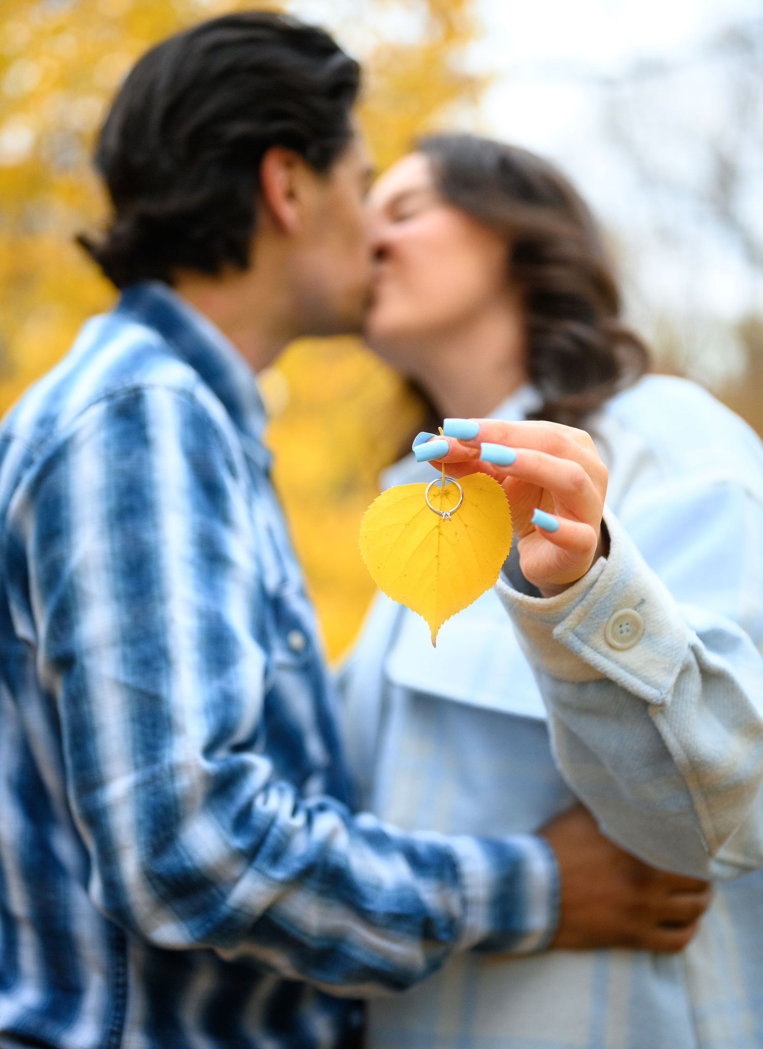 A couple shares a kiss while holding a yellow leaf against a backdrop of autumn colors.