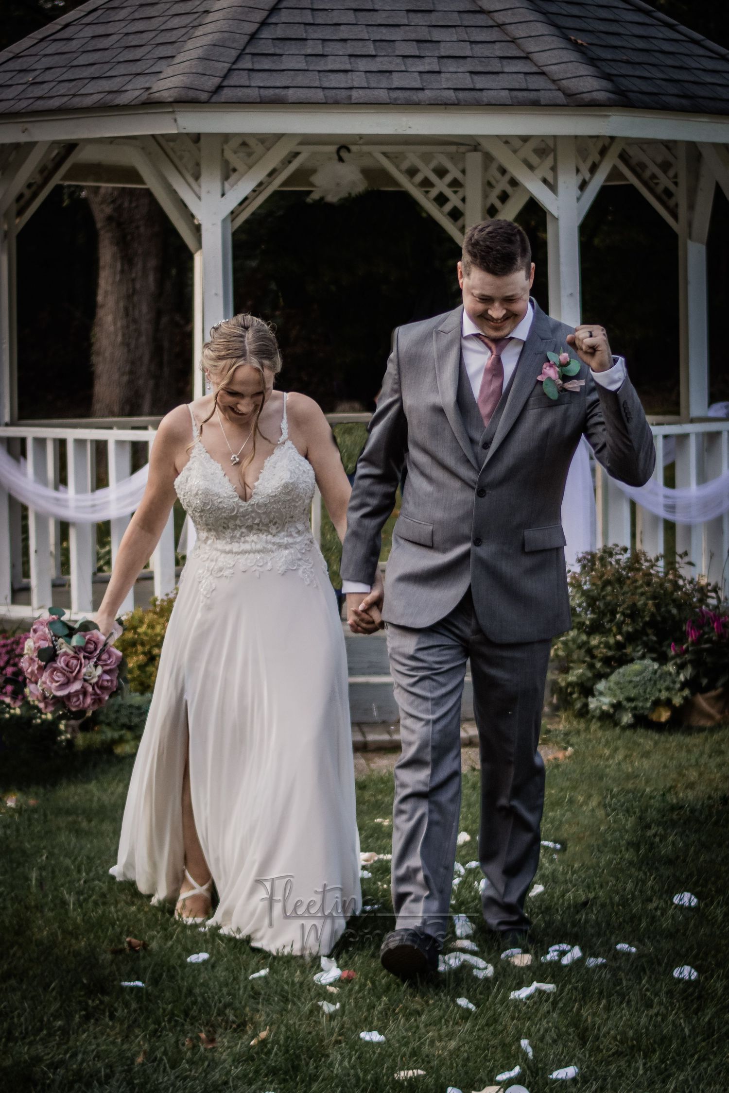 Newlyweds walking hand in hand near gazebo with flower petals scattered on ground.
