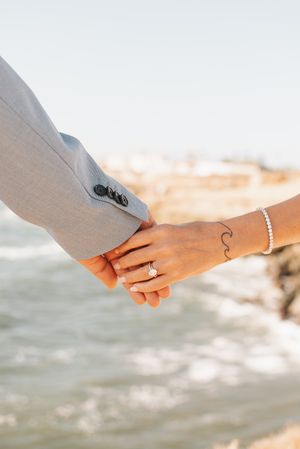 Close-up of couple's hands with wave tattoo and wedding rings against ocean background.