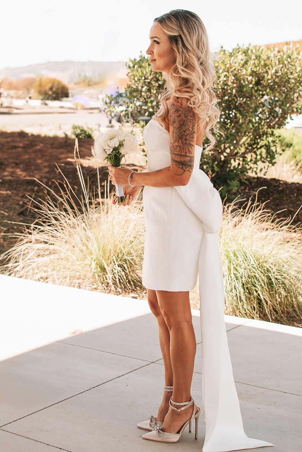 Modern bridal look featuring short dress with dramatic bow train and silver heels.