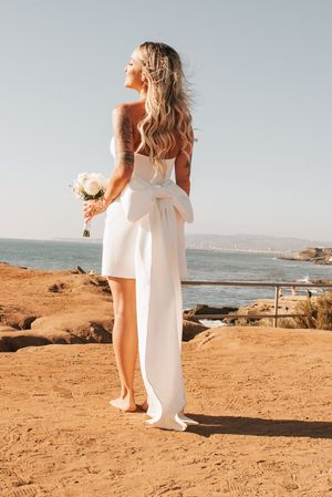 Bride in white dress with bow detail standing on coastal cliff overlooking ocean.