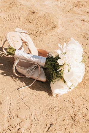 Elegant bridal shoes and white bouquet arranged on sandy beach with soft lighting.