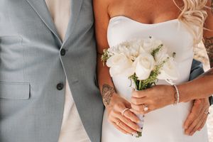 Wedding couple detail showing white dress, gray suit, and floral bouquet with visible tattoo artwork.