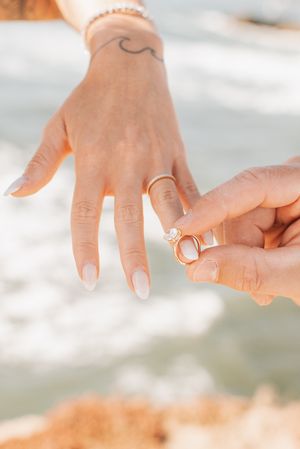 Close-up of engagement ring being placed on finger with wave tattoo and ocean backdrop.