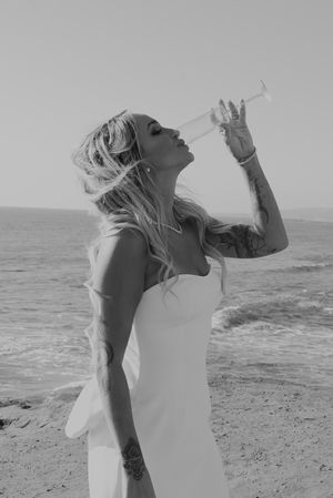 Woman in white dress drinking from champagne glass by ocean.