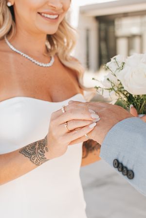 Close-up of wedding couple's hands with diamond ring and white bouquet, featuring elegant necklace and lace dress details.