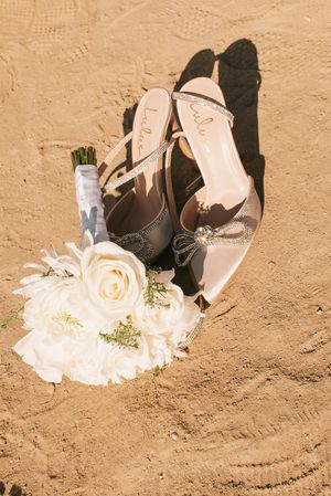 White bridal heels and floral bouquet with roses casting shadows on beach sand.