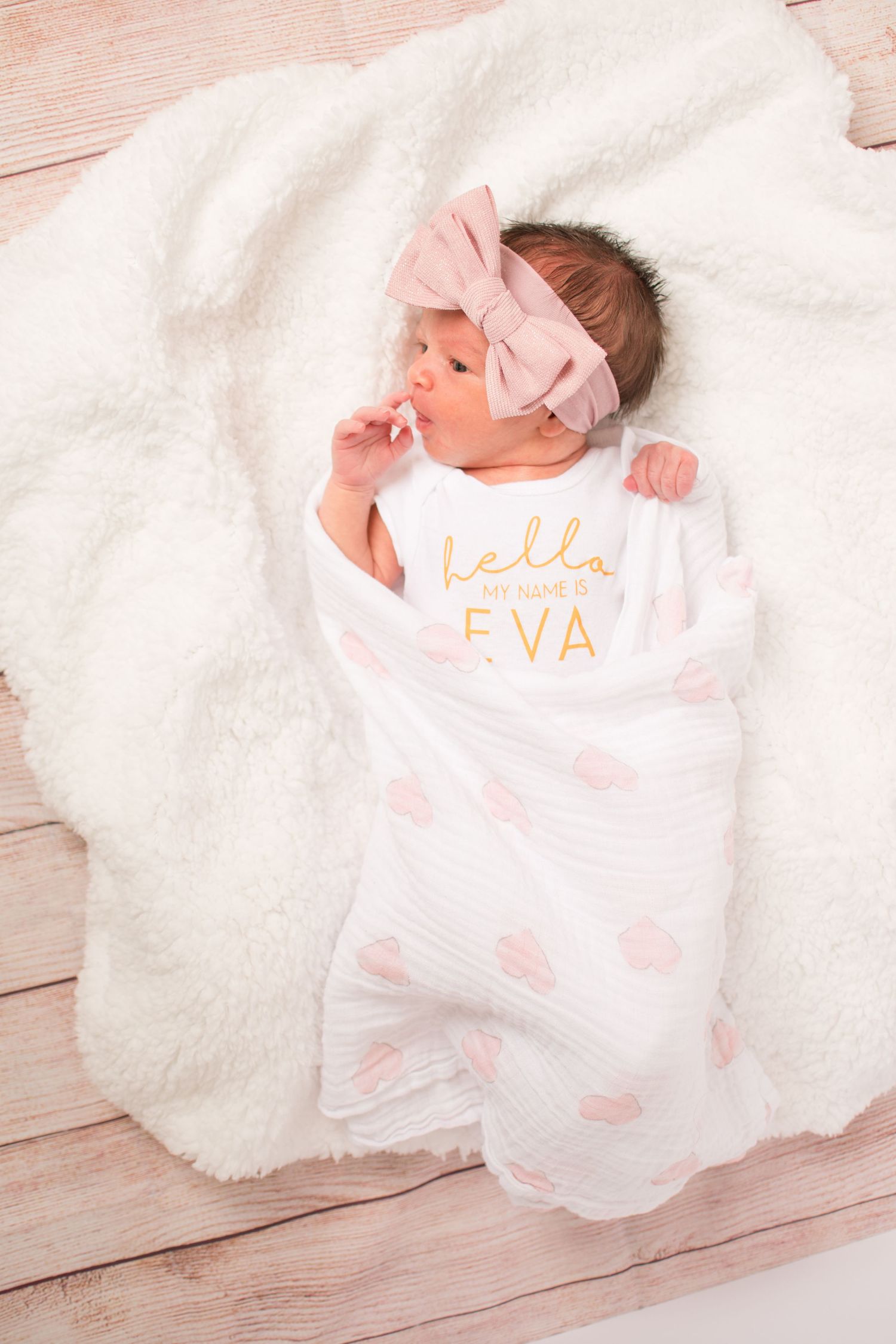 Baby girl laying swaddled on a fuzzy blanket. She has a big pink bow on and her shirt says hello.