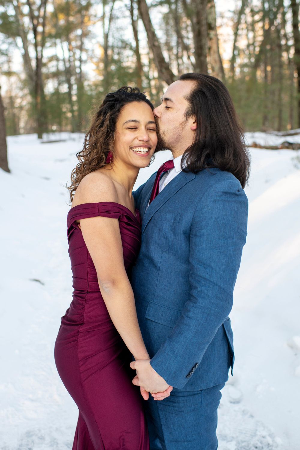 Man kissing his fiancée on the cheek as they hold hands during a snowy Massachusetts engagement session at Walden Pond
