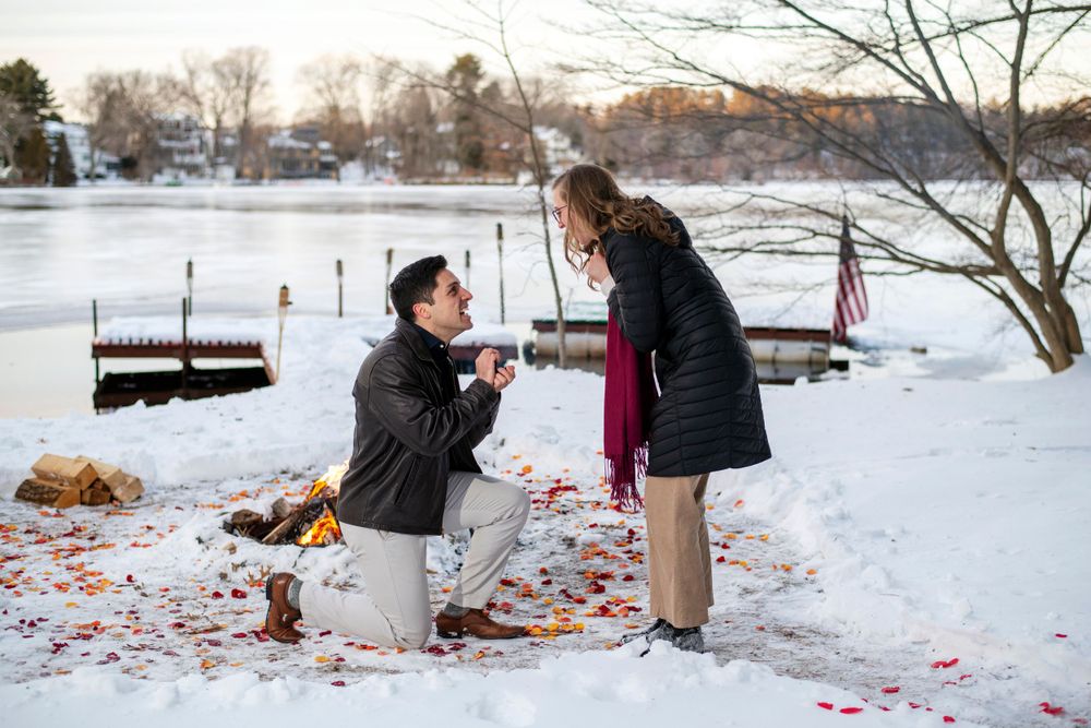 Surprise winter proposal beside a frozen lake, with the groom-to-be kneeling in the snow with an engagement ring