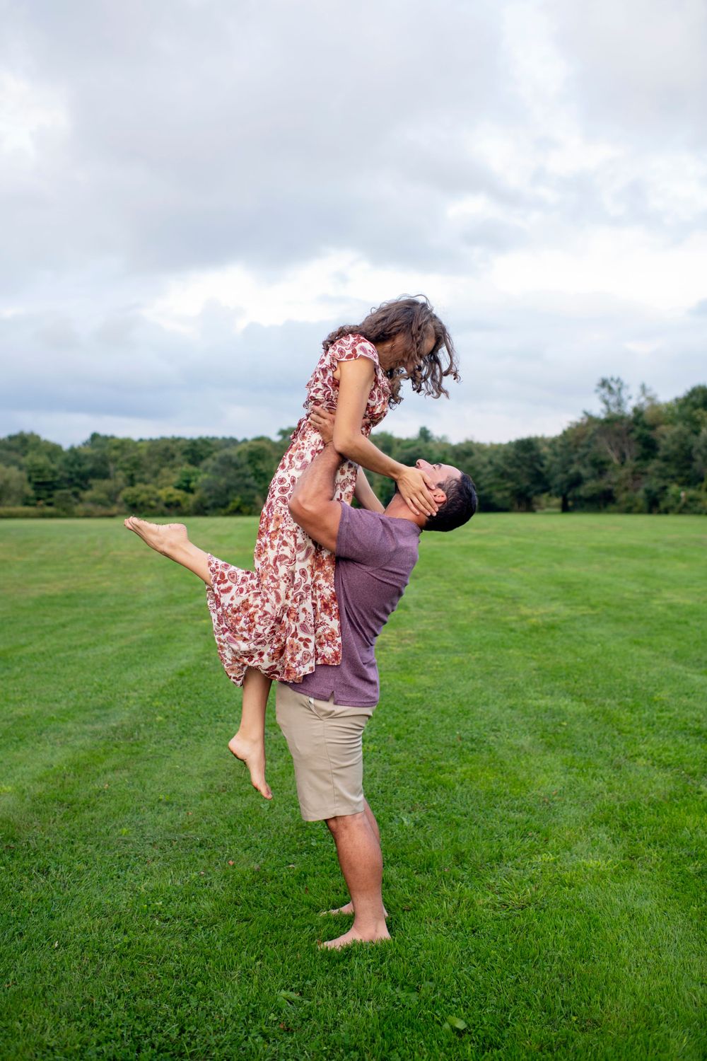 Man lifting his fiancée into the air during a playful outdoor engagement session in a green field