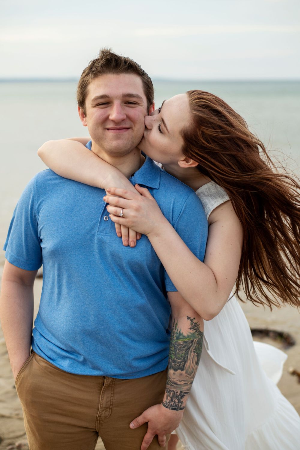 Woman kissing her fiancé on the cheek while embracing him during a breezy New England beach engagement session