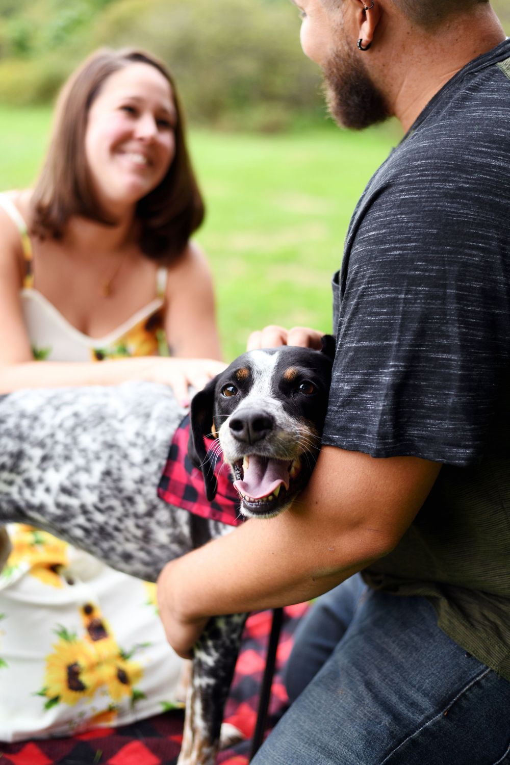 Happy dog wearing a red plaid bandana with its engaged owners during an outdoor engagement session