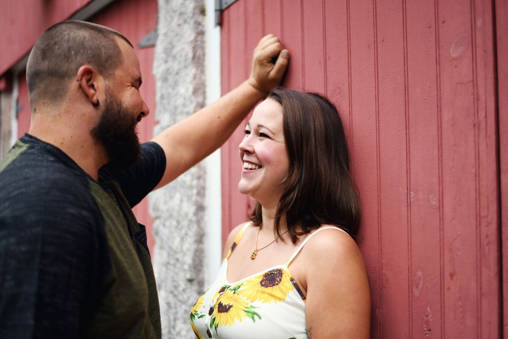 Couple laughing together beside a rustic red barn during a casual outdoor engagement session