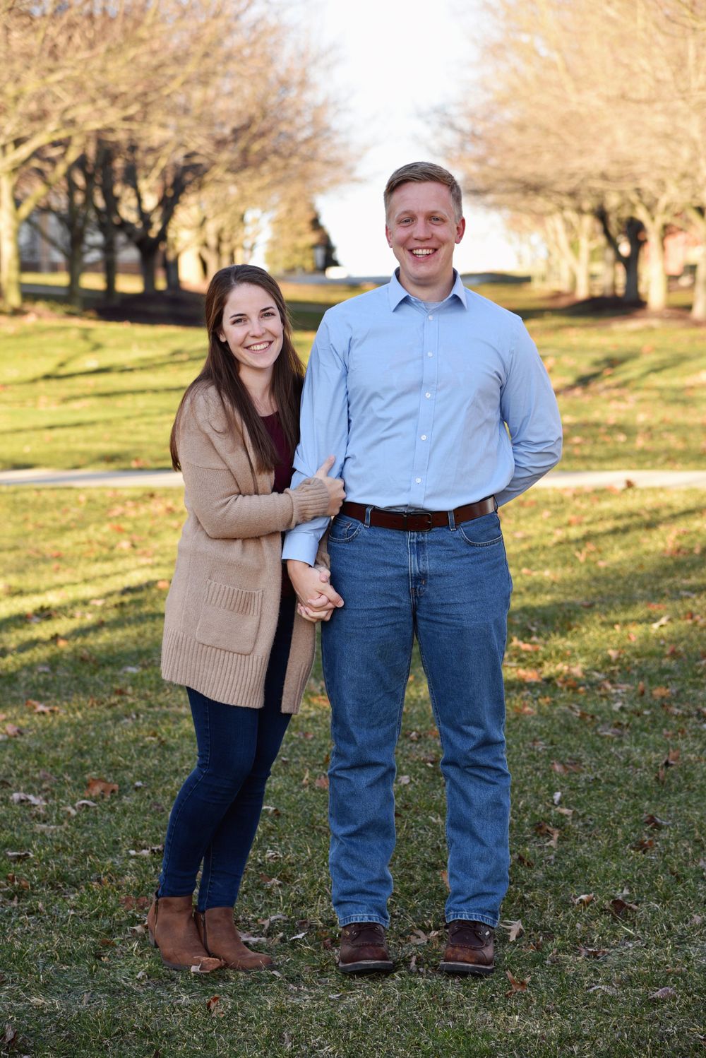 Smiling engaged couple holding hands beneath a tree-lined path during a fall engagement session at Franciscan University