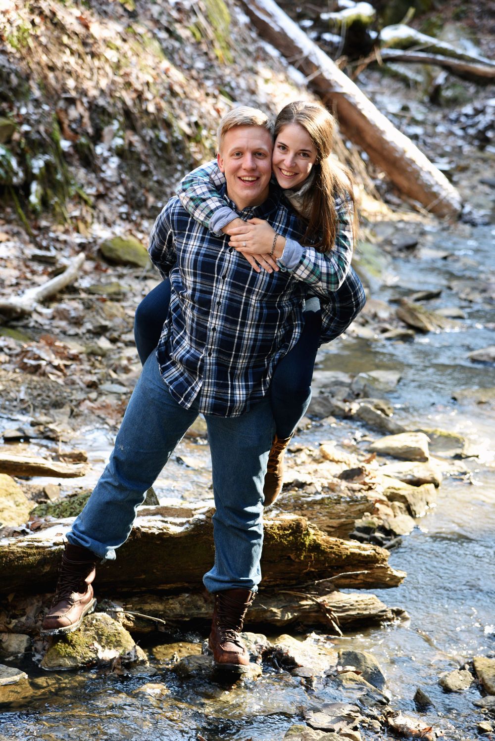Man giving his fiancé a piggyback ride while crossing a shallow creek during a playful outdoor engagement session