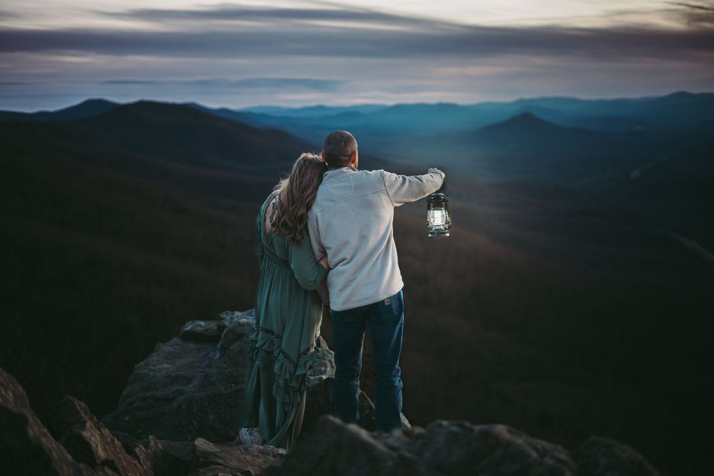A couple sits together on a mountain overlook during blue hour with distant peaks.