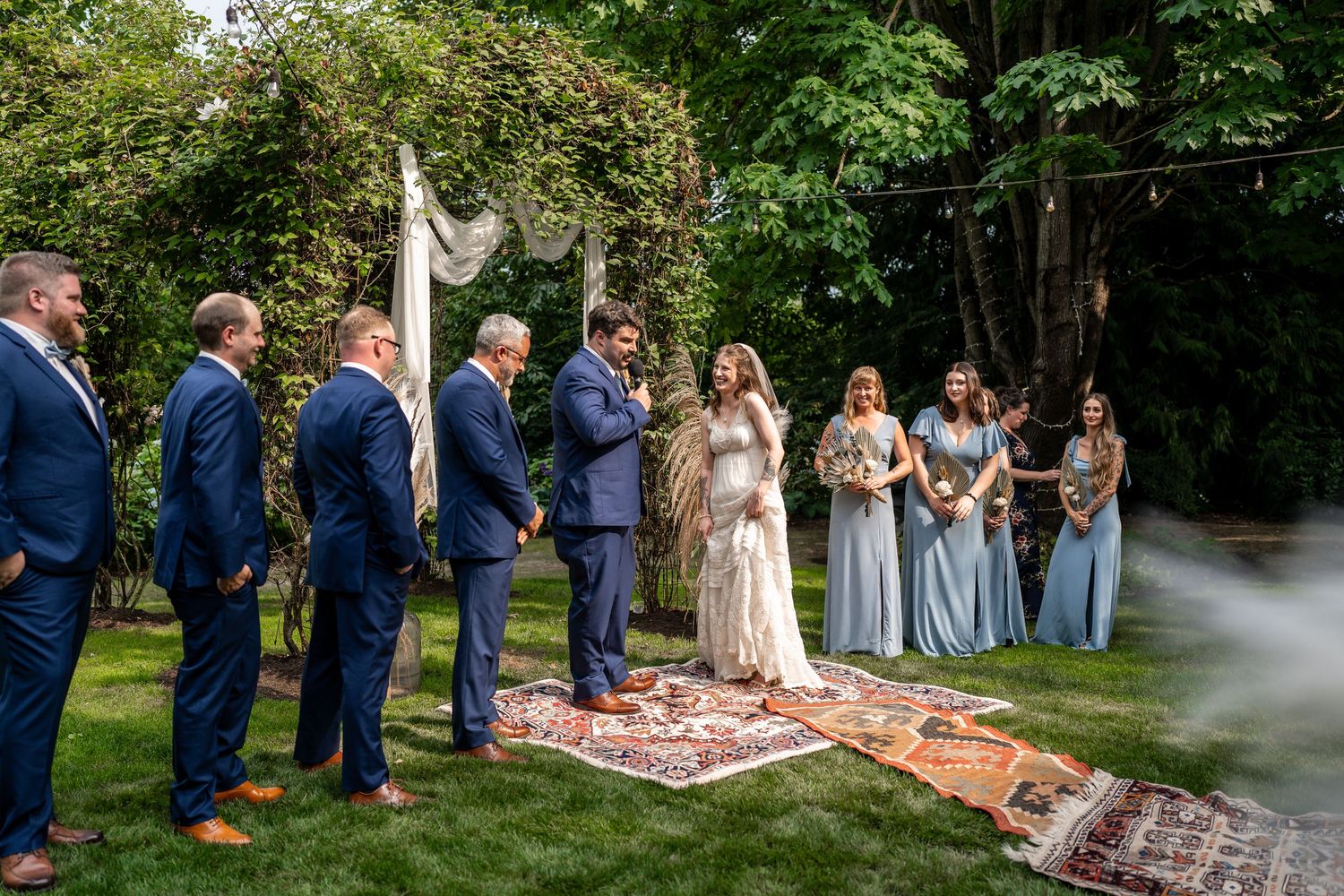 Wedding ceremony outdoors with wedding party in blue and sage dresses standing on vintage rugs.