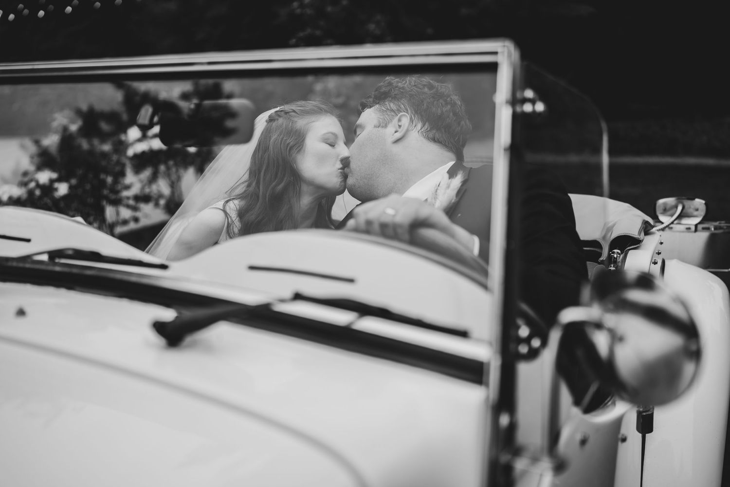 Black and white romantic photo of newlyweds sharing a kiss in vintage convertible car.