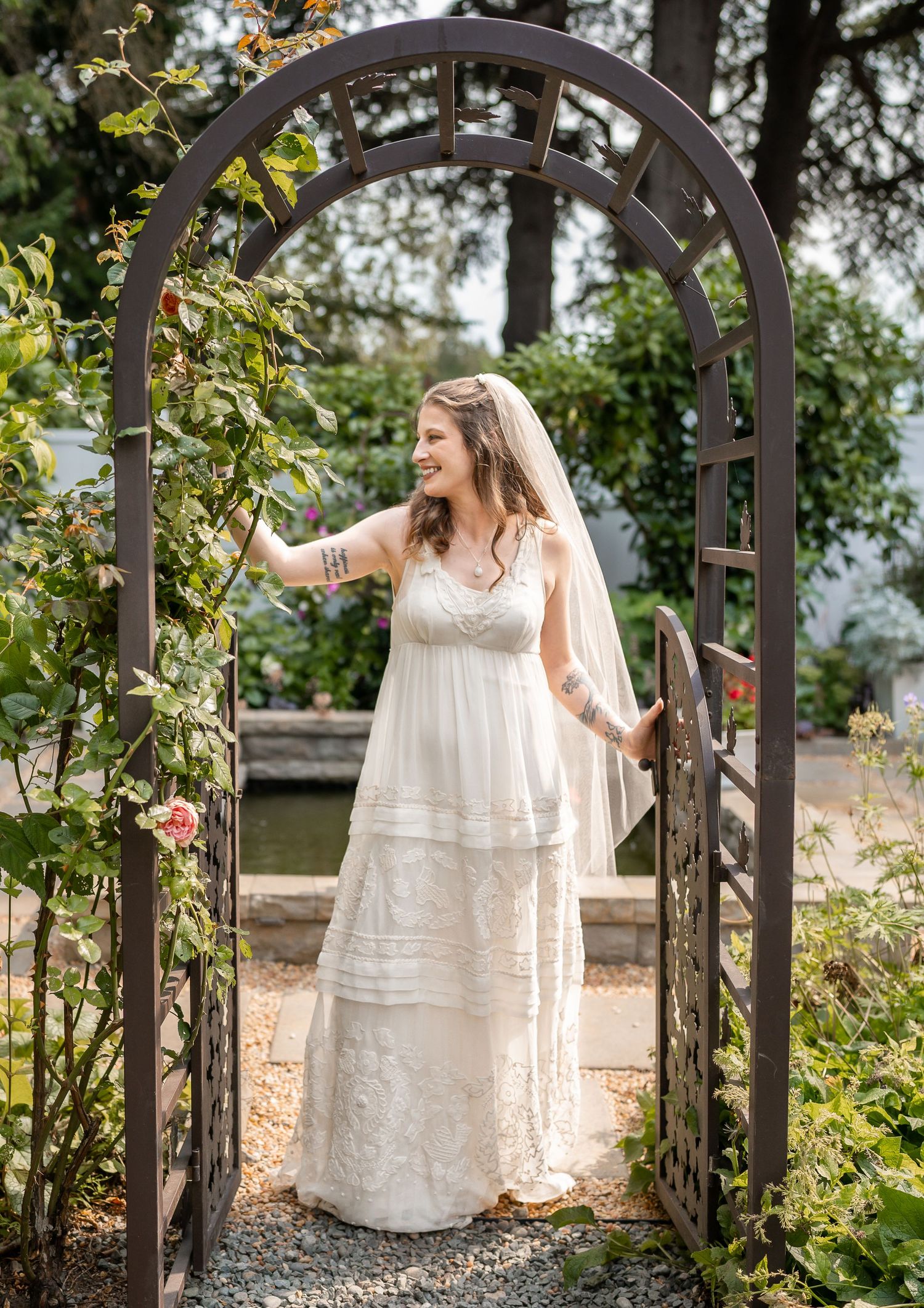 A bride in a flowing lace wedding dress stands under a garden archway surrounded by greenery and roses.