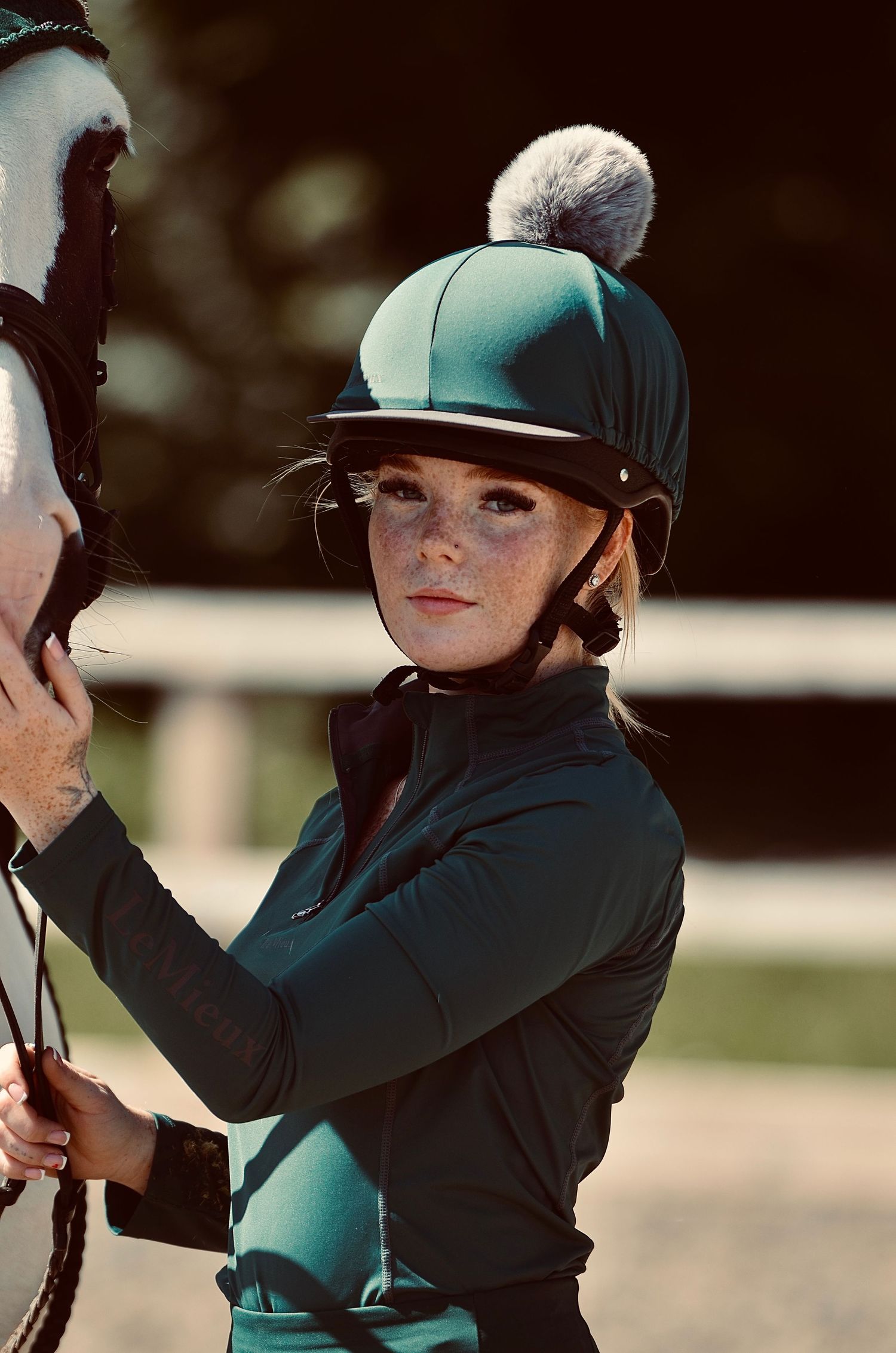 Young female equestrian with freckles wearing a teal riding helmet with pom-pom standing beside a white horse.