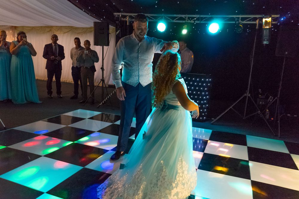 Newlyweds dance on a checkered floor under blue and purple lighting in a reception tent.