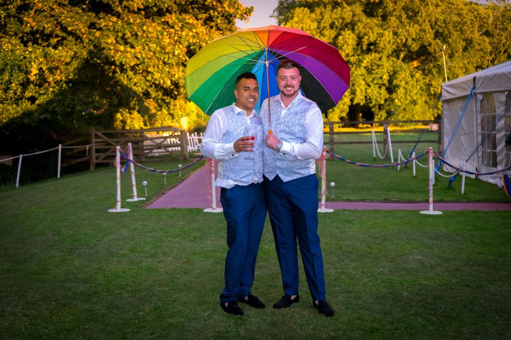A couple stands beneath a rainbow umbrella on their wedding day in an outdoor setting.