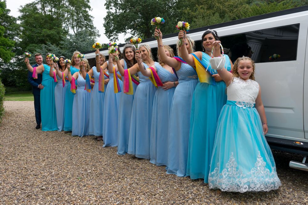 Bridesmaids in blue dresses pose enthusiastically beside a white limousine.