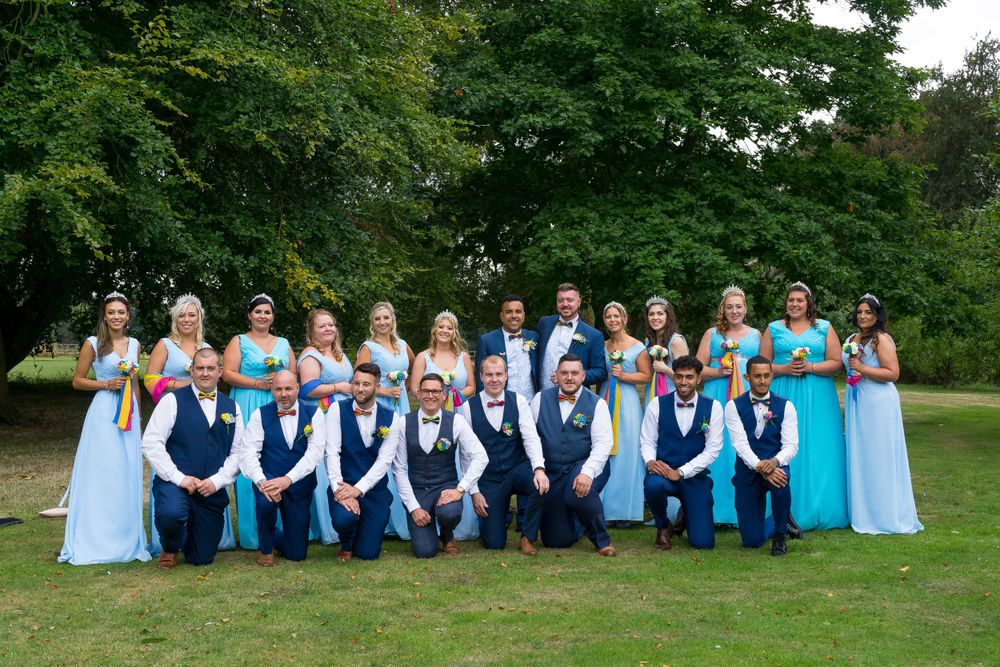Wedding party in blue dresses and navy vests pose together on a green lawn.