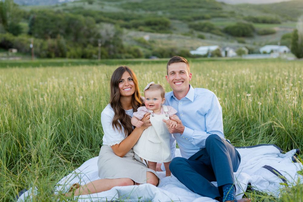 Happy family of three with baby girl sitting on a blanket in a green field with mountain views at sunset.