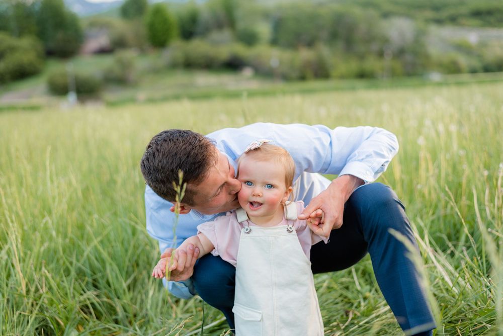 Father kissing his laughing toddler daughter in a lush green field during a sunny family photo session.