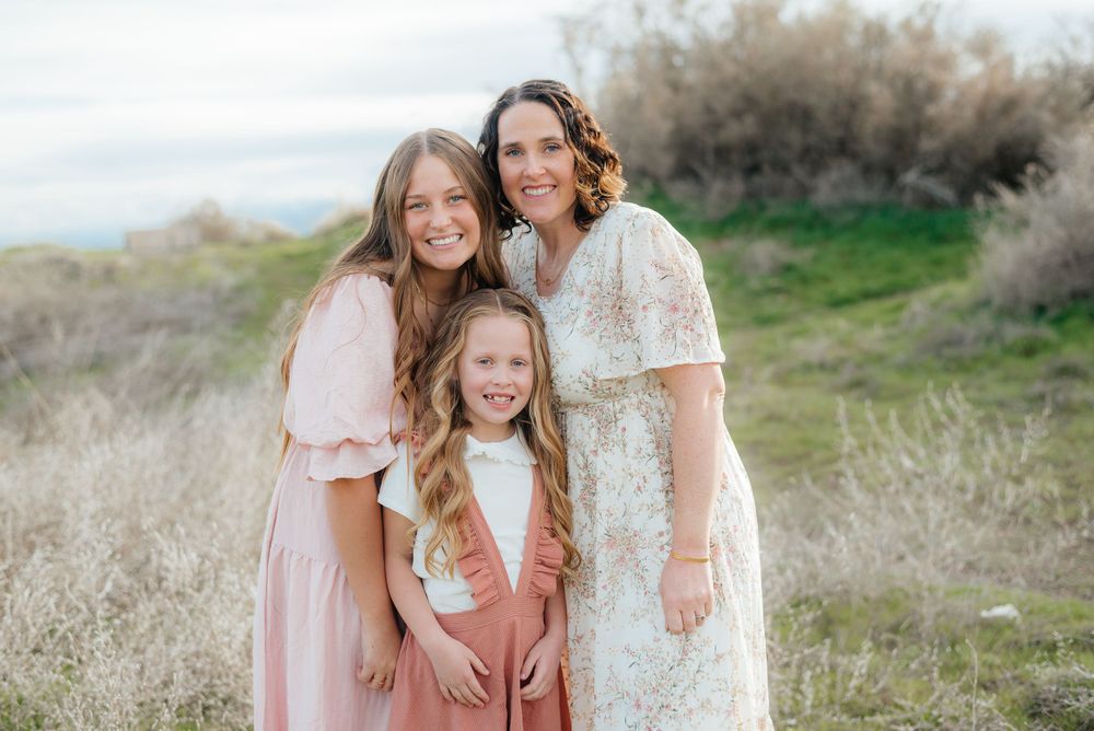 Mother poses warmly with two daughters in a sunlit outdoor field.